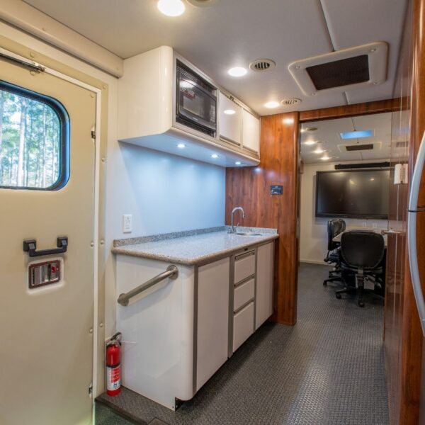 Interior shot of a clean, modern command center kitchenette with a granite counter, microwave, sink, and safety door leading into the main mobile conference room.